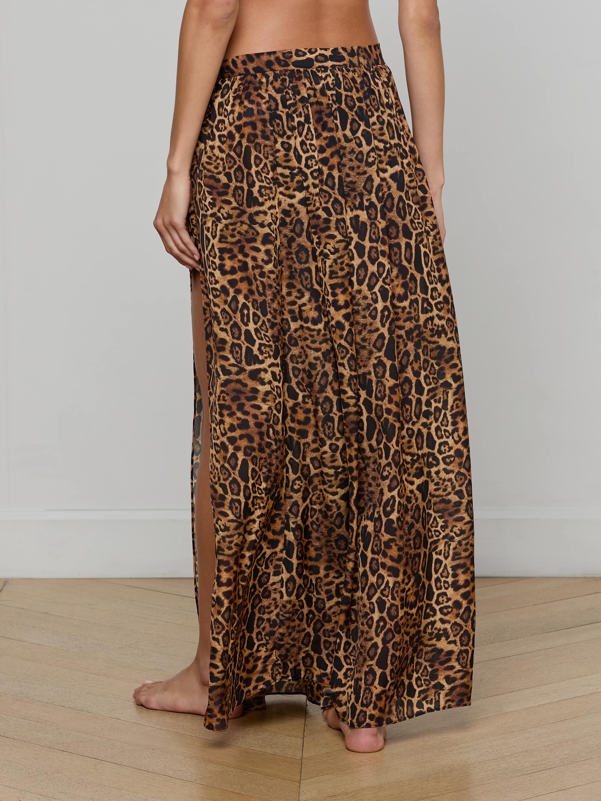 A woman stands barefoot, shown from the back against a plain light background, wearing the Klaire Cover-Up Skirt—a long, high-slit wrap skirt crafted from viscose chiffon and featuring a brown and black leopard print pattern.