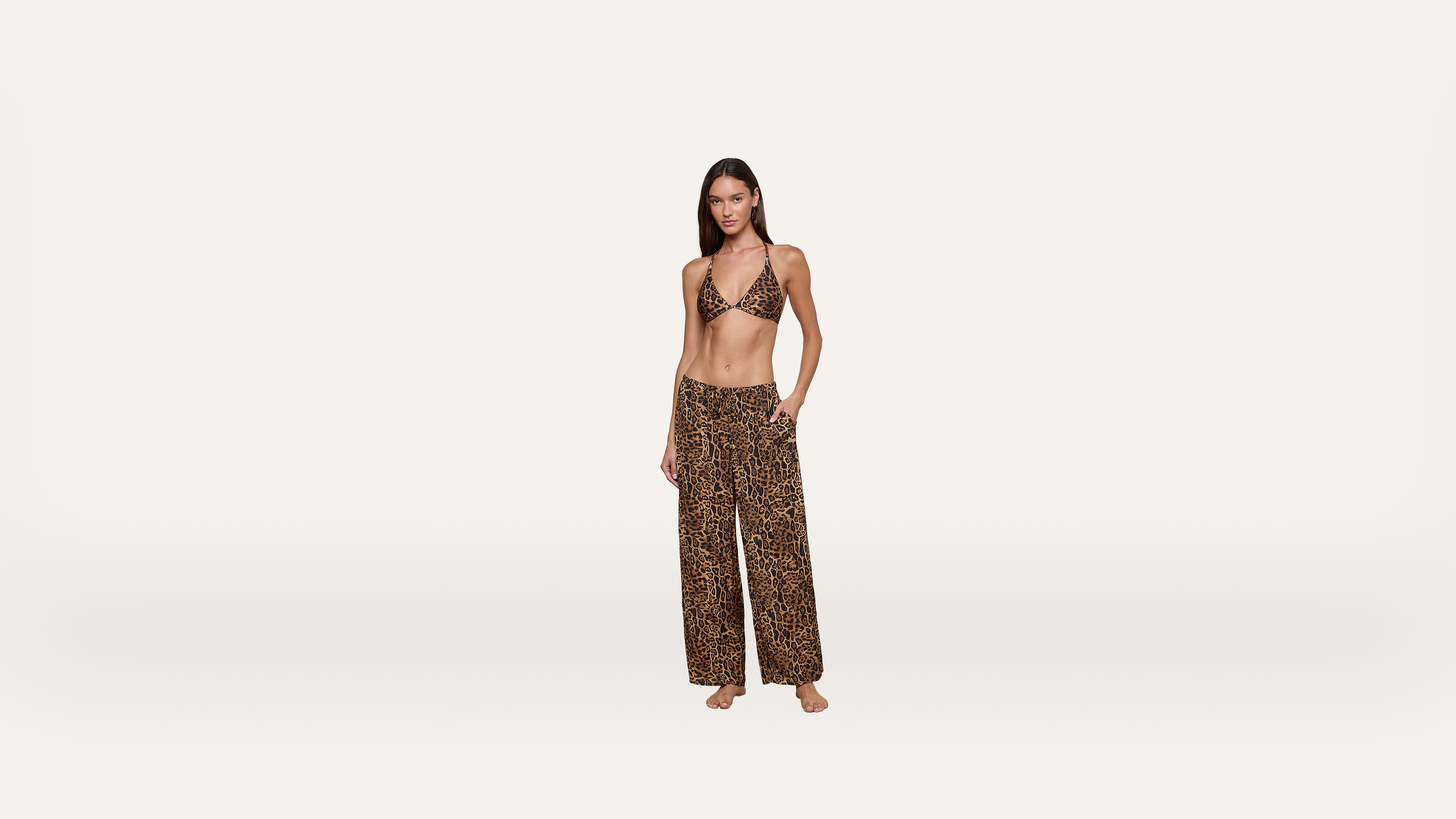A woman stands barefoot wearing a leopard print bikini top and matching wide-leg pants against a plain white background.