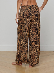 A person stands barefoot, viewed from the back, wearing the Geraldine Cover-Up Pant—a wide-leg, leopard print style with a relaxed pull-on fit. The upper body is mostly out of frame.