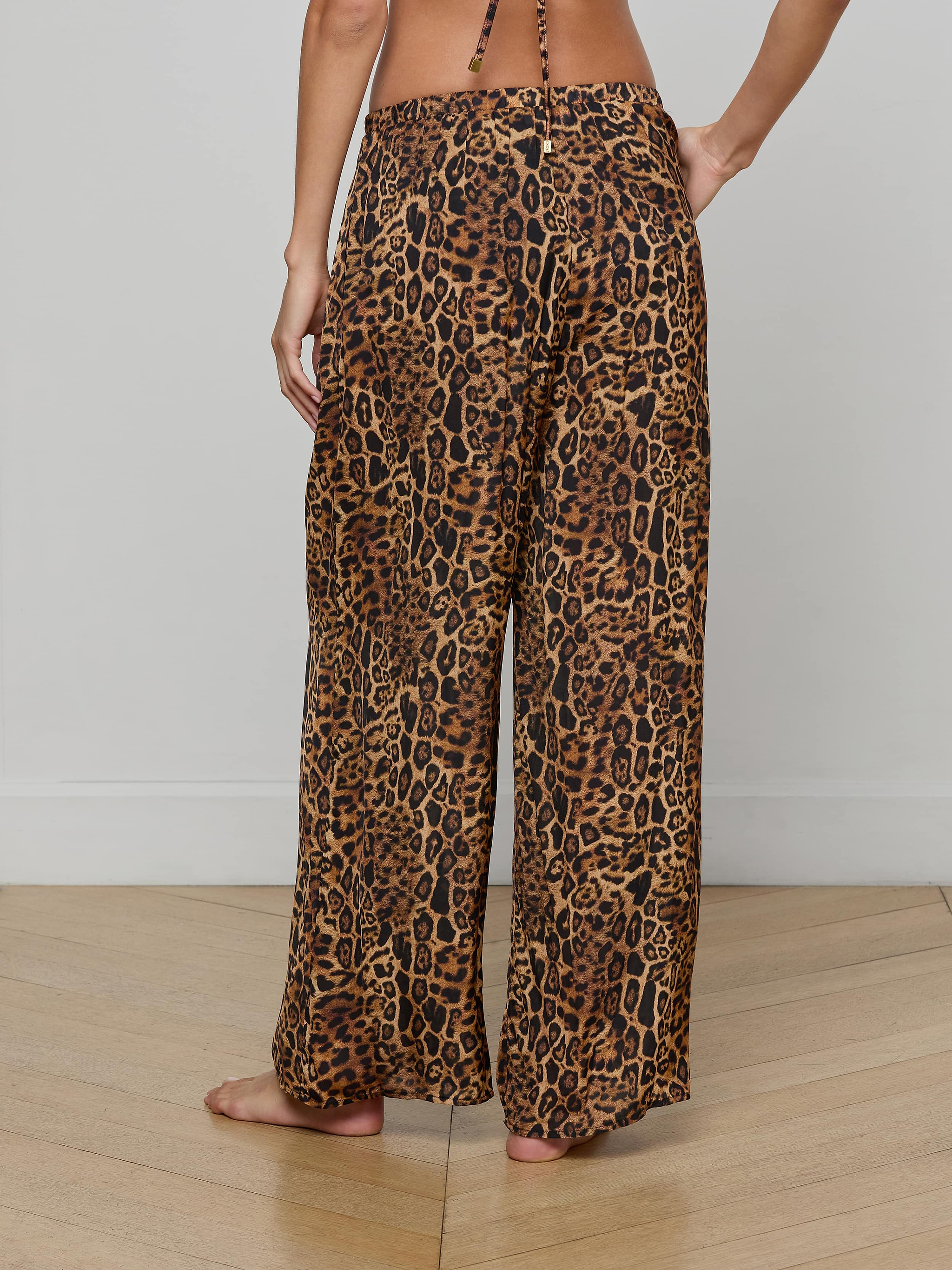 A person stands barefoot, viewed from the back, wearing the Geraldine Cover-Up Pant—a wide-leg, leopard print style with a relaxed pull-on fit. The upper body is mostly out of frame.