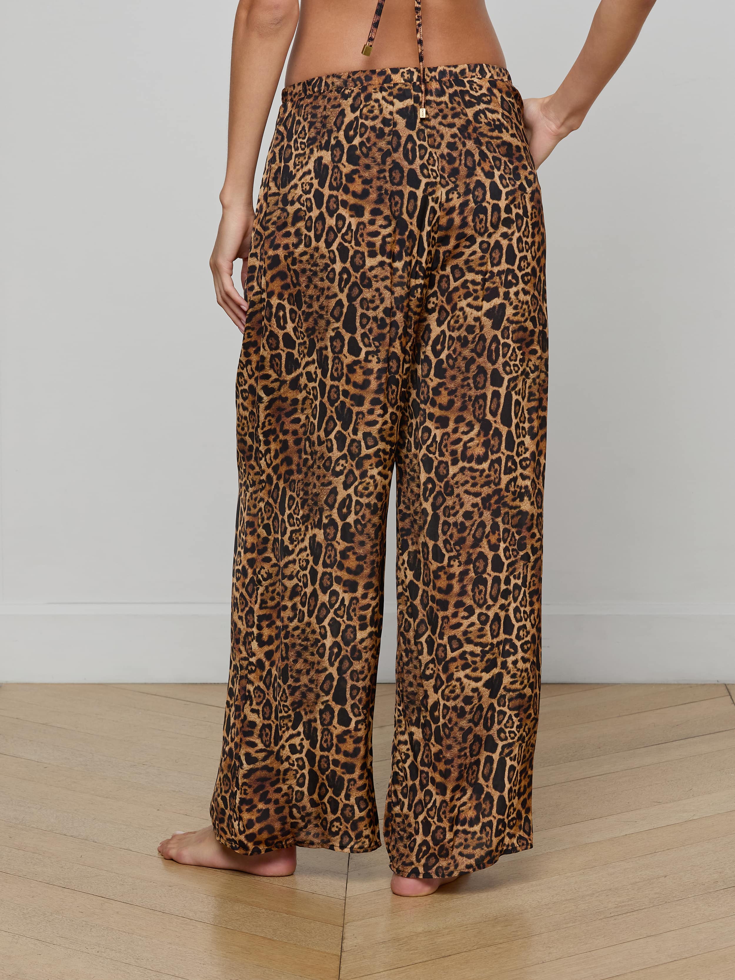 A person stands barefoot, viewed from the back, wearing the Geraldine Cover-Up Pant—a wide-leg, leopard print style with a relaxed pull-on fit. The upper body is mostly out of frame.