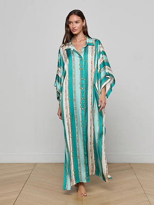 A woman faces forward on a wooden floor in the L'AGENCE Wrena Cover-Up Dress—a long, teal and white striped button-down kaftan beach dress with wide sleeves and gold accents—showcasing effortless resort style against a light gray wall.