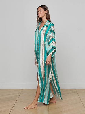 A woman stands barefoot on a wooden floor, wearing the L'AGENCE Wrena Cover-Up Dress—a loose, long teal and white striped kaftan with gold accents—perfect as a beach dress or cover-up. She faces left against a plain light gray wall.