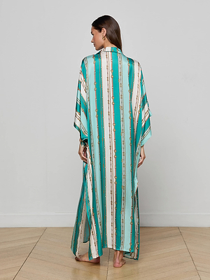 A woman stands barefoot on a wooden floor, facing away from the camera, wearing the L'AGENCE Wrena Cover-Up Dress—a long, flowing teal resort piece with vertical gold and white patterns—against a plain light gray wall.
