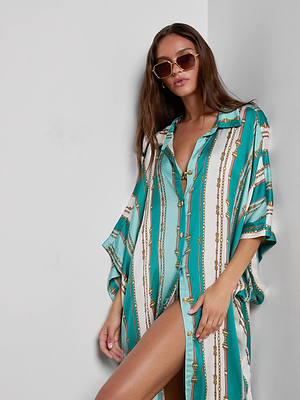 A woman with long brown hair wears sunglasses and the L'AGENCE Wrena Cover-Up Dress, a loose turquoise and white striped shirt dress with gold accents, standing against a white wall and looking at the camera.