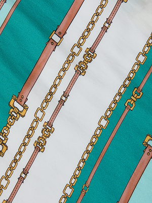 The Marceline Short Pareo features a lightweight viscose rayon fabric with a teal and white belt-stripe print, accented by brown belts and gold chains—ideal for a chic, sage-inspired look.