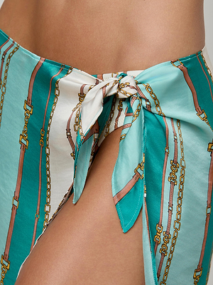 Close-up of a person wearing the Marceline Short Pareo—a turquoise and white wrap skirt with gold chain and belt patterns, tied at the hip as a stylish swimsuit cover-up, revealing part of their upper thigh.