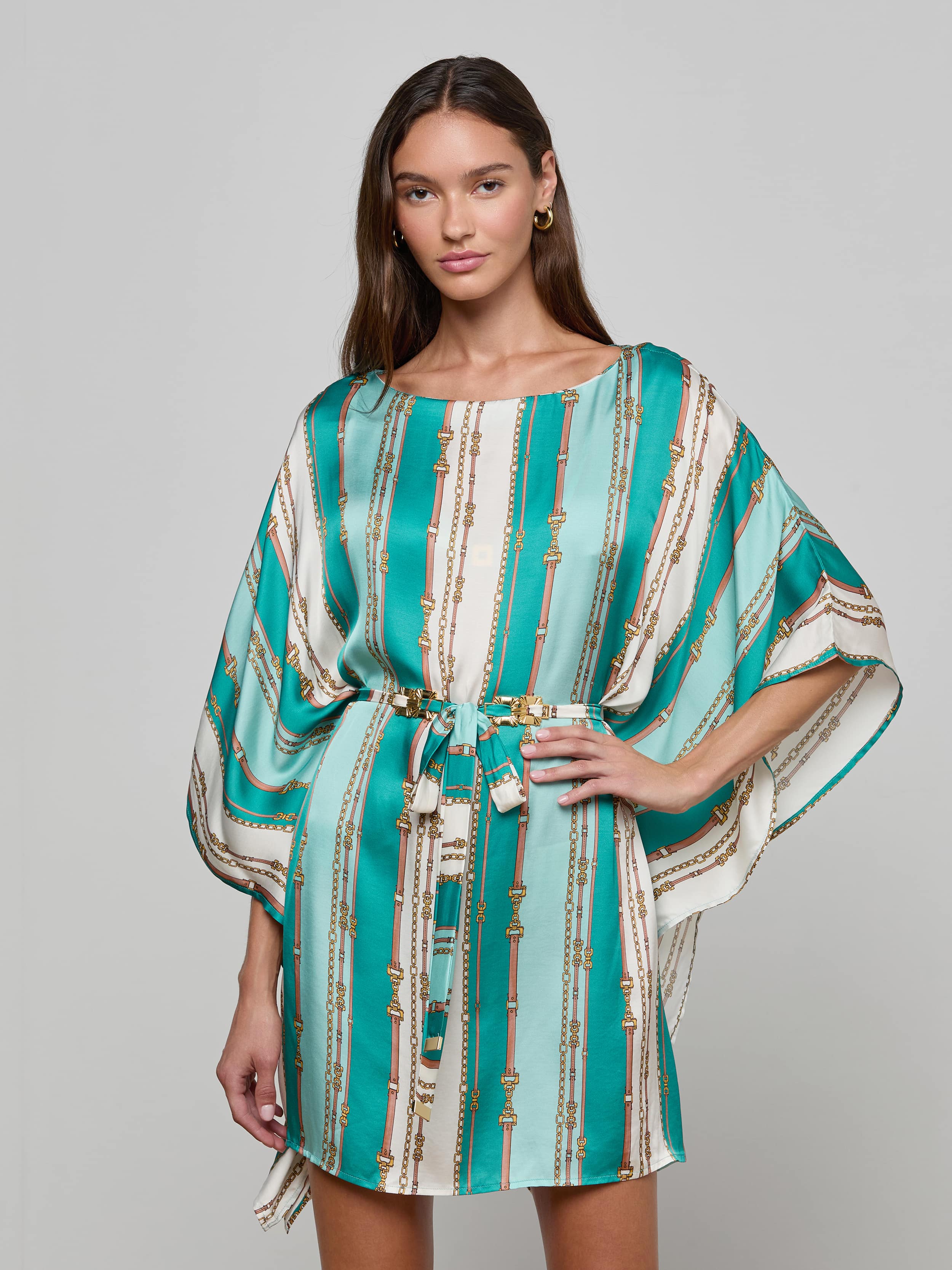 A woman wearing the Alia Cover-Up Tunic in a short, turquoise and white pattern with gold accents, wide sleeves, and a fabric belt at the waist stands against a plain background.