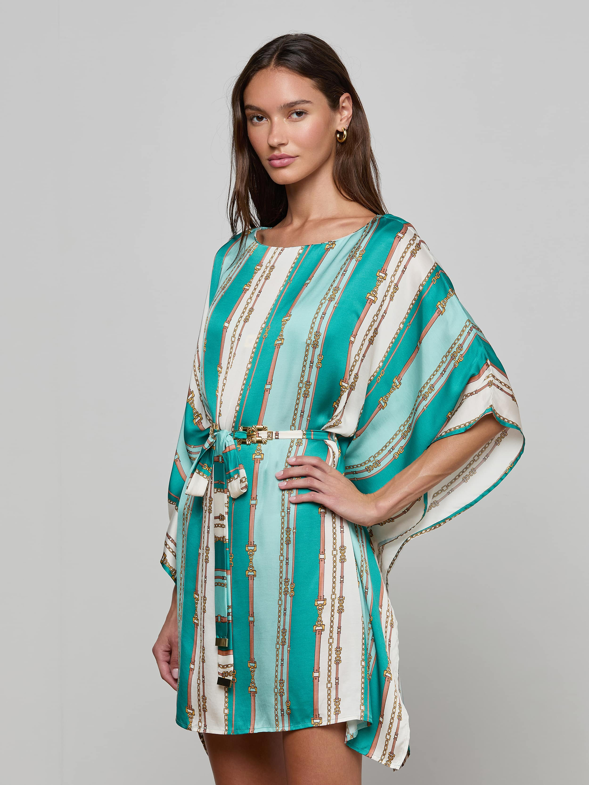 Woman wearing the Alia Cover-Up Tunic, featuring a turquoise and white belt-stripe print with gold accents, standing against a plain background.