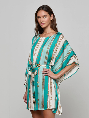 A woman models the Alia Cover-Up Tunic, featuring a turquoise and white belt-stripe print with gold chain details, a belted waist, and loose sleeves; she stands against a plain background, looking at the camera with one hand on her hip.