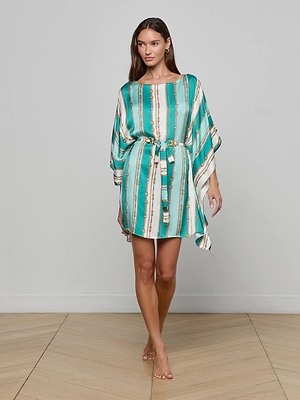 A woman stands indoors facing forward, smiling slightly as she wears the Alia Cover-Up Tunic in turquoise and white belt-stripe print with gold accents and a tied waist. She is barefoot on a light wooden floor against a plain gray wall.