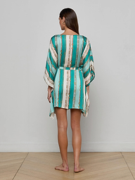A woman stands barefoot facing away against a plain white background, wearing the Alia Cover-Up Tunic—a short, loose design with long sleeves and a turquoise and white belt-stripe print.