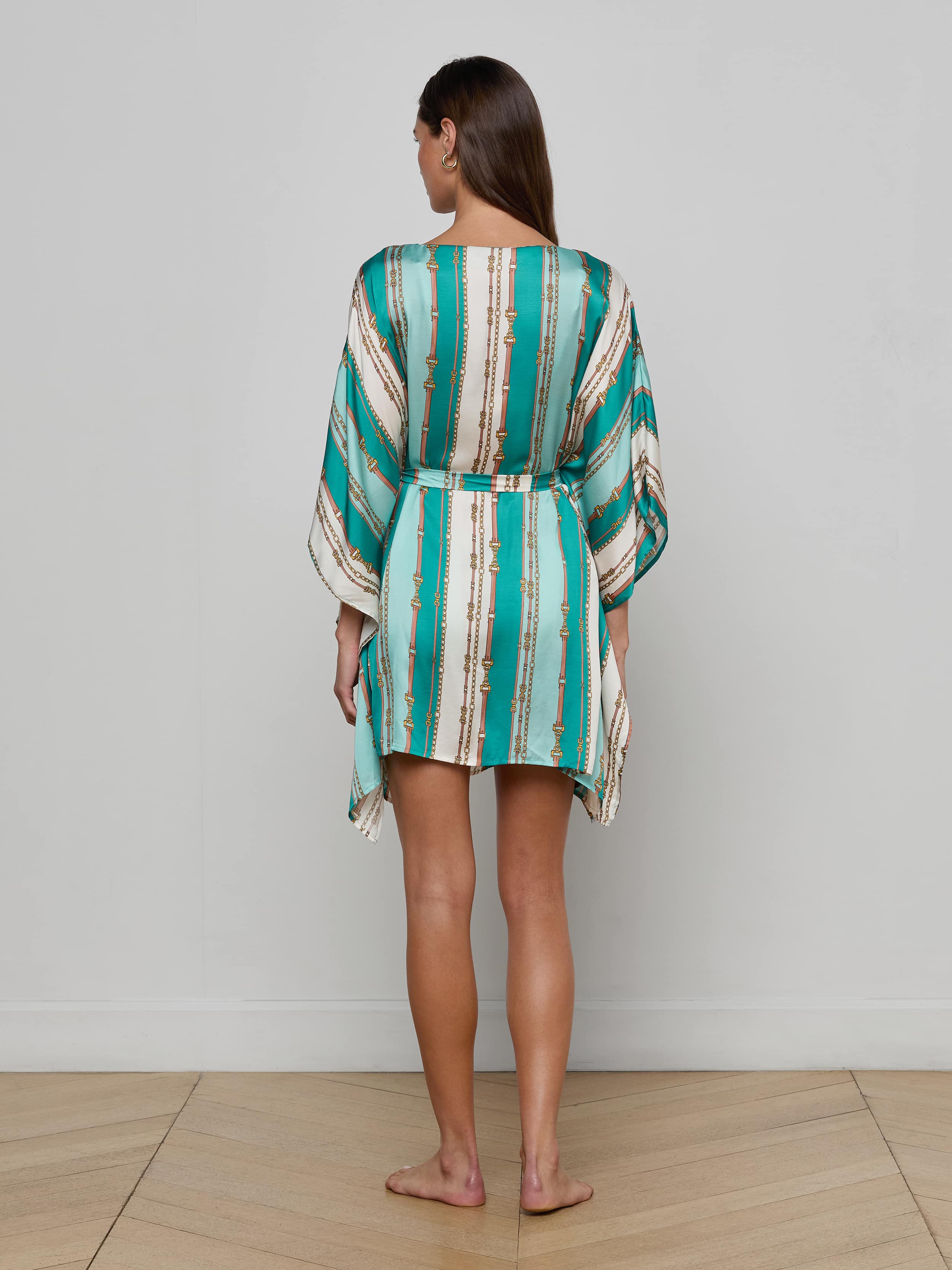 A woman stands barefoot facing away against a plain white background, wearing the Alia Cover-Up Tunic—a short, loose design with long sleeves and a turquoise and white belt-stripe print.