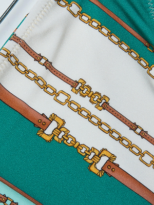 Close-up of the Annabelle Triangle Bikini Top fabric, featuring brown belts and gold chains on a white and teal background. Detailed buckles and stitching create a luxurious, stylish swimwear design.