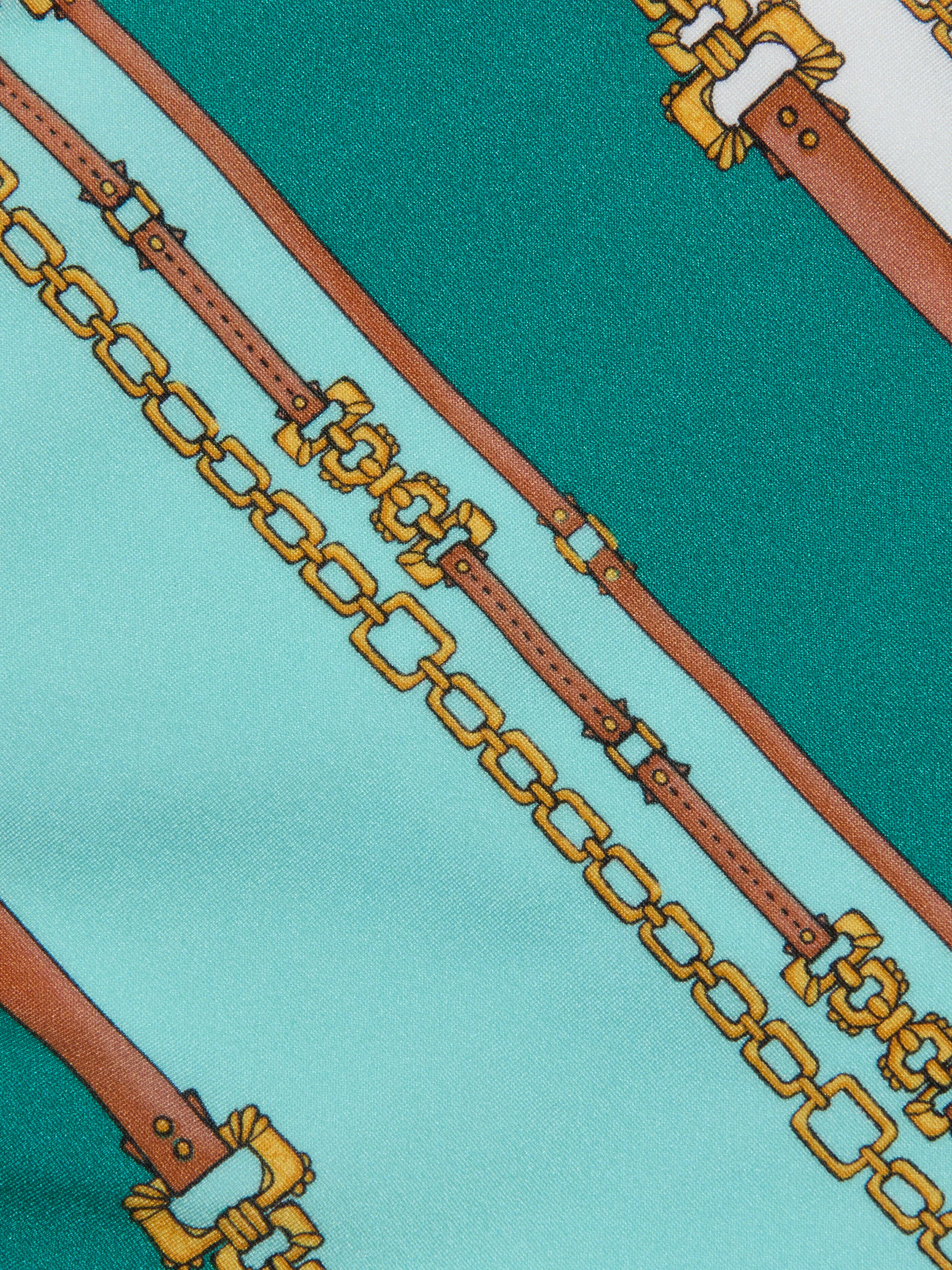 Nicole Bikini Bottom in a patterned fabric with teal and light blue stripes, featuring a belt-stripe print accented by brown leather straps and gold chains running diagonally across the surface.