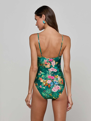 A woman with long brown hair faces away, wearing the Lilou One-Piece Swimsuit in green, featuring a high-leg cut and a vibrant floral-butterfly print. She stands against a plain light gray background, hands by her sides.