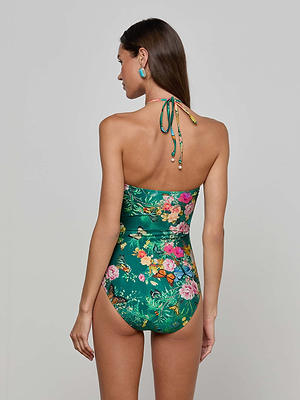 A woman with long brown hair and large green earrings stands with her back to the camera, wearing the Shiva One-Piece Swimsuit featuring a green butterfly floral print and a halter neck tie against a plain light background.