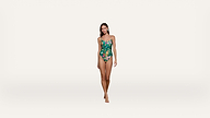 A woman stands barefoot against a plain white background, wearing a green floral one-piece swimsuit with thin straps.