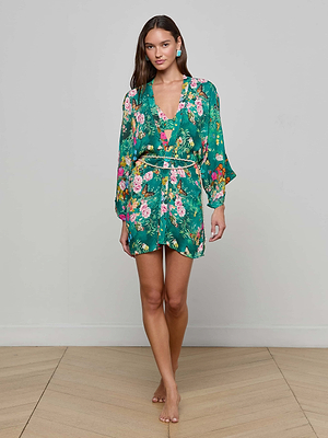 A woman stands barefoot on a wooden floor wearing the Caterina Cover-Up Kimono, a green floral wrap with long sleeves, a removable belt, tied waist, and delicate crochet trim, against a plain light gray wall.