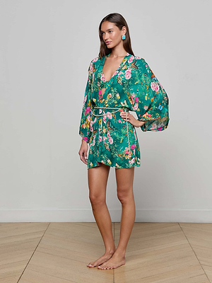 A woman stands barefoot on a wooden floor, wearing the green Caterina Cover-Up Kimono with long, wide sleeves and a removable belt. She has long brown hair, turquoise earrings, and is posed against a plain light gray wall.