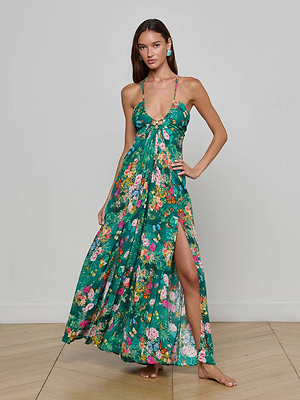 A woman in the Geneva Cover-Up Dress—a green floral maxi with a deep V-neck, spaghetti straps, and dual high slits—stands barefoot on a wooden floor against a plain light gray wall.