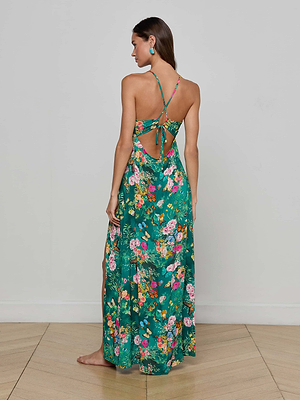 A woman with long brown hair wears the Geneva Cover-Up Dress, a green floral halter style with an open back and tie detail. She stands barefoot on a wood floor, facing away from the camera against a plain white wall—ideal for resort evenings.