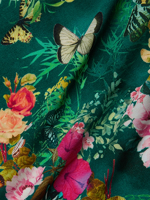 A close-up of fabric with a vibrant floral and butterfly print on green, featuring pink flowers, yellow and white butterflies, plus green foliage. The Caterina Cover-Up Kimono includes a removable belt and delicate floral crochet trim.