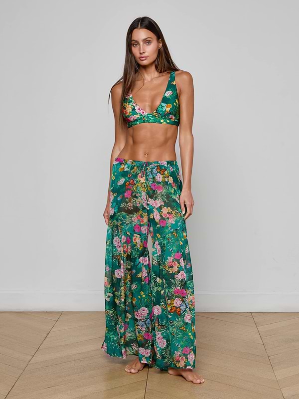 A woman stands barefoot against a plain white background, wearing the Sharon Cover-Up Pant—a chic green floral long skirt with a drawstring waist—paired with a matching bikini top. This stylish beachwear look is perfect for warm, sunny days.