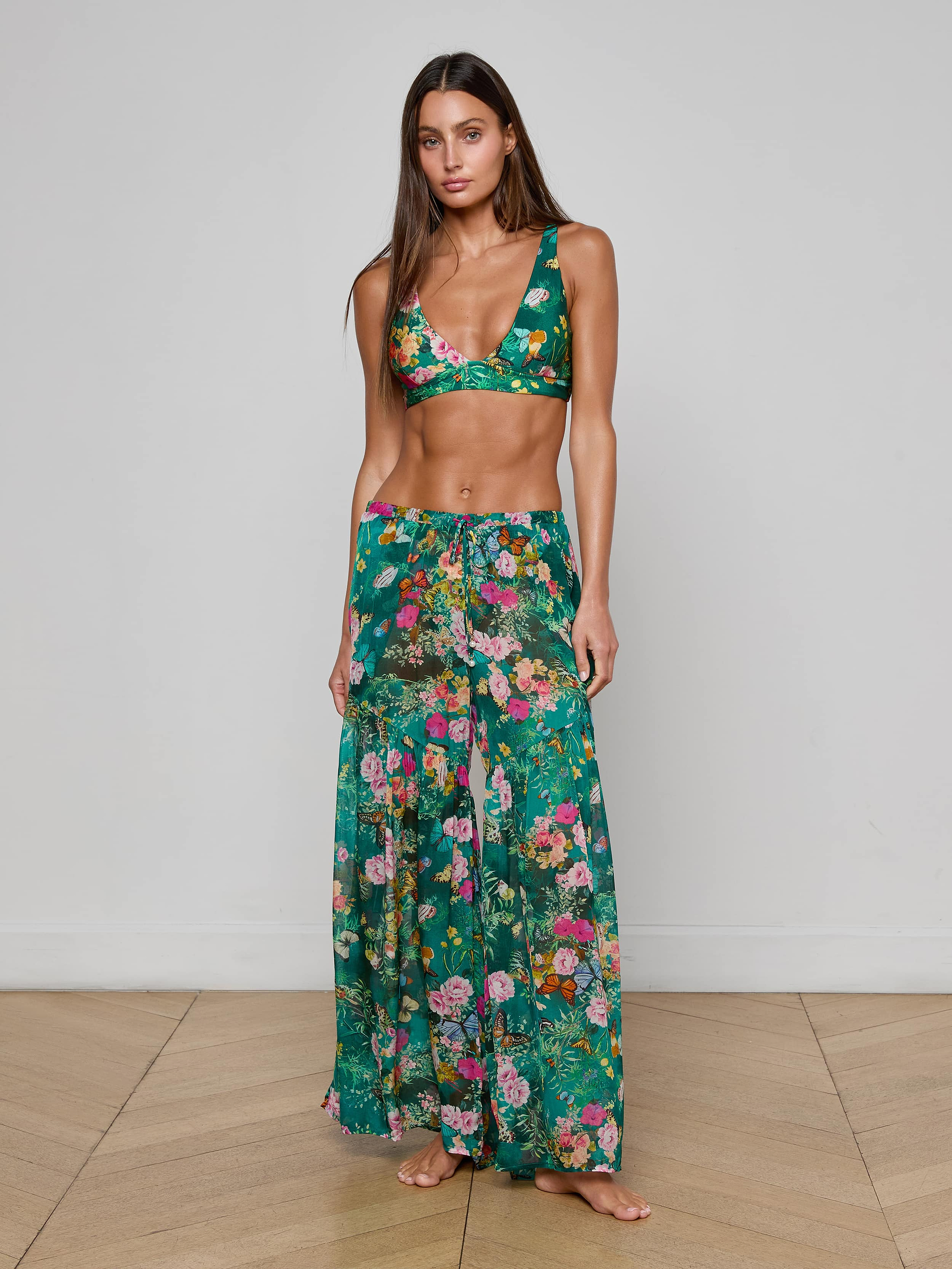 A woman stands barefoot against a plain white background, wearing the Sharon Cover-Up Pant—a chic green floral long skirt with a drawstring waist—paired with a matching bikini top. This stylish beachwear look is perfect for warm, sunny days.