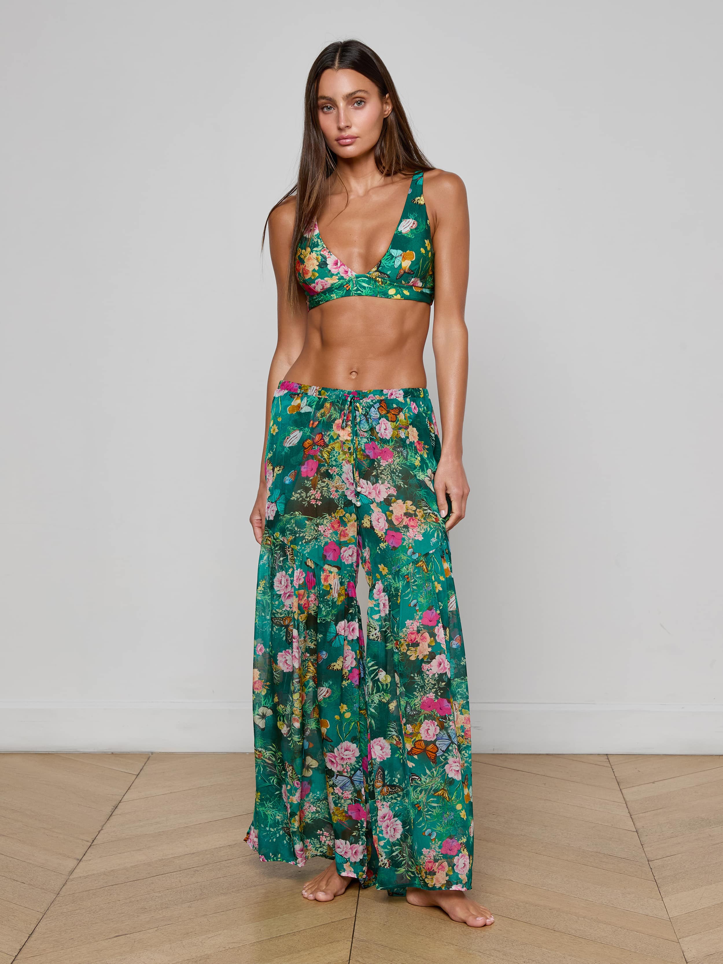 A woman stands barefoot against a plain white background, wearing the Sharon Cover-Up Pant—a chic green floral long skirt with a drawstring waist—paired with a matching bikini top. This stylish beachwear look is perfect for warm, sunny days.