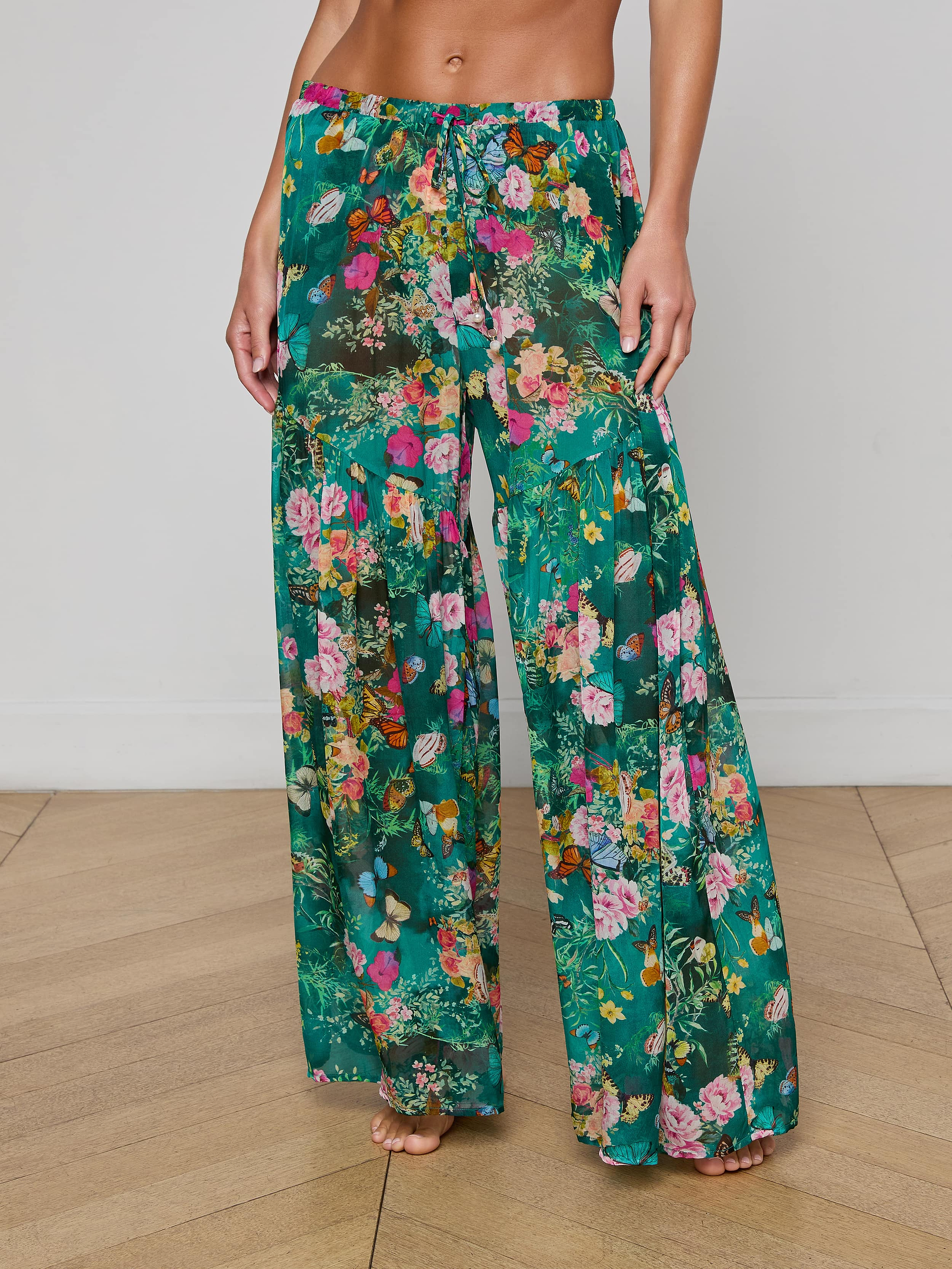 A person stands barefoot against a plain background, showcasing the lower torso and legs while wearing the Sharon Cover-Up Pant—loose, wide-leg green floral pants featuring a drawstring waist.