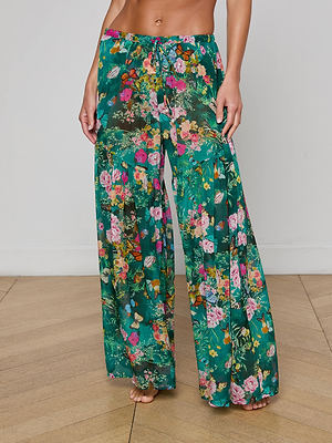 A person stands barefoot on a wooden floor wearing L'AGENCE Sharon Cover-Up Pant—loose, high-waisted green drawstring pants with a colorful floral print. Their upper body is not fully visible.