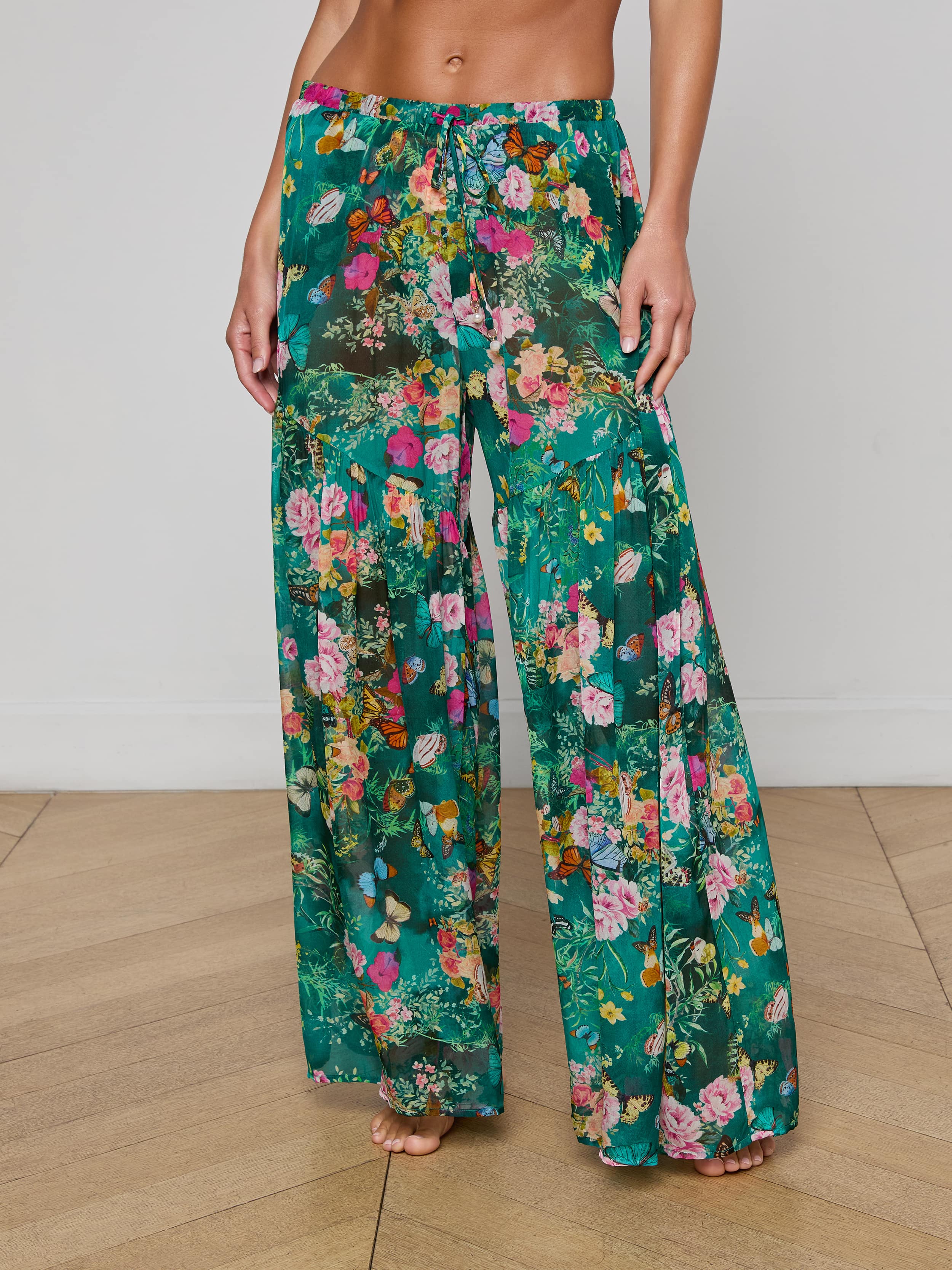 A person stands barefoot against a plain background, showcasing the lower torso and legs while wearing the Sharon Cover-Up Pant—loose, wide-leg green floral pants featuring a drawstring waist.