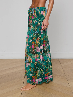 A woman stands barefoot on a wooden floor, with only her lower torso and legs visible, wearing the Sharon Cover-Up Pant by L'AGENCE—a green floral maxi skirt with pink and yellow flowers and a drawstring waist, ideal for stylish beachwear.