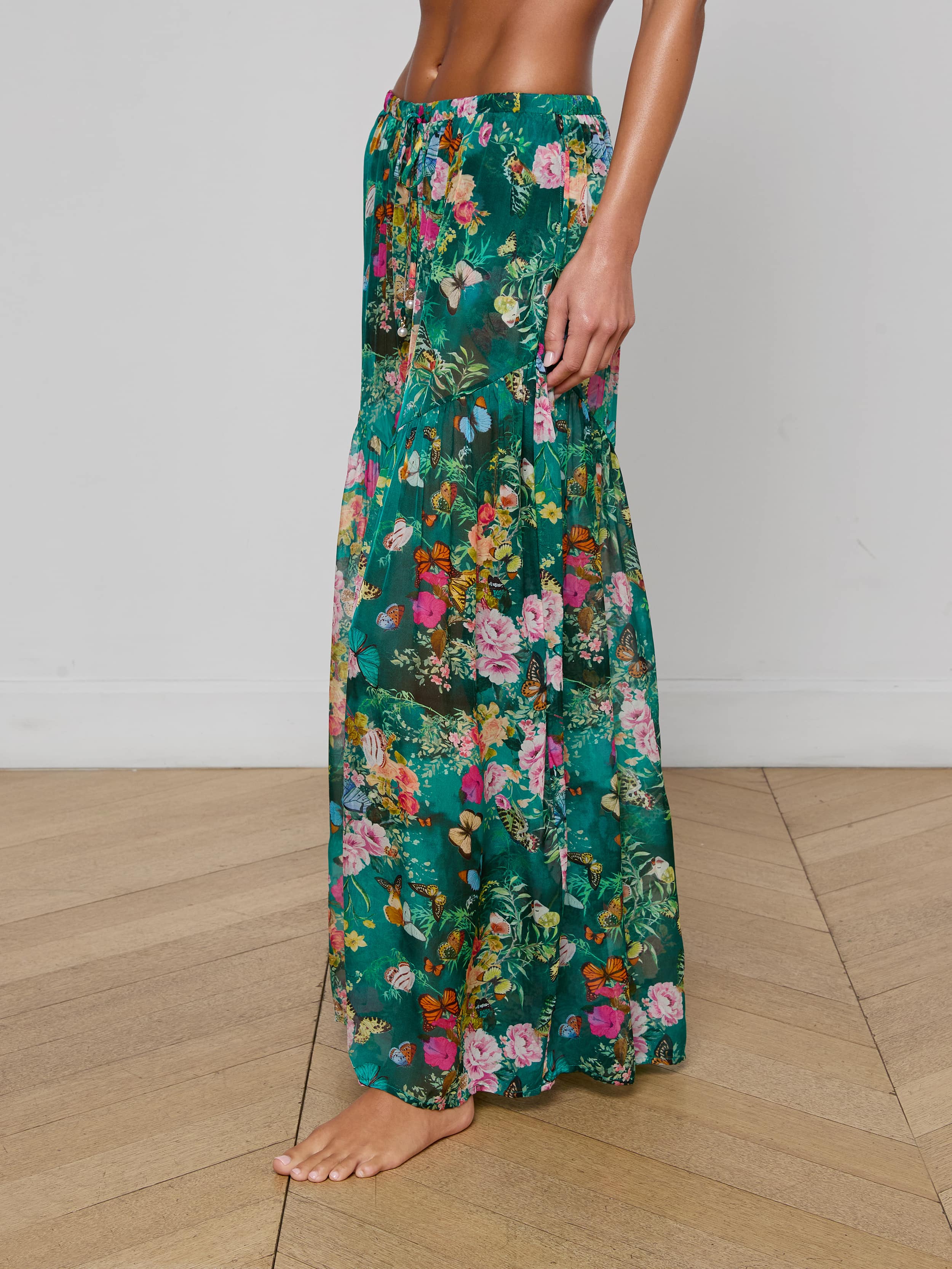 A woman stands barefoot, wearing the Sharon Cover-Up Pant—a long, green floral design with a drawstring waist. Her upper body is cropped out, and the background is plain white.