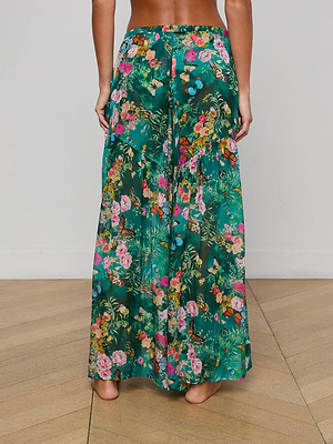 A woman stands barefoot on a wooden floor, seen from behind wearing the Sharon Cover-Up Pant by L'AGENCE—a long, flowy green pant with a drawstring waist and vibrant floral and butterfly print.