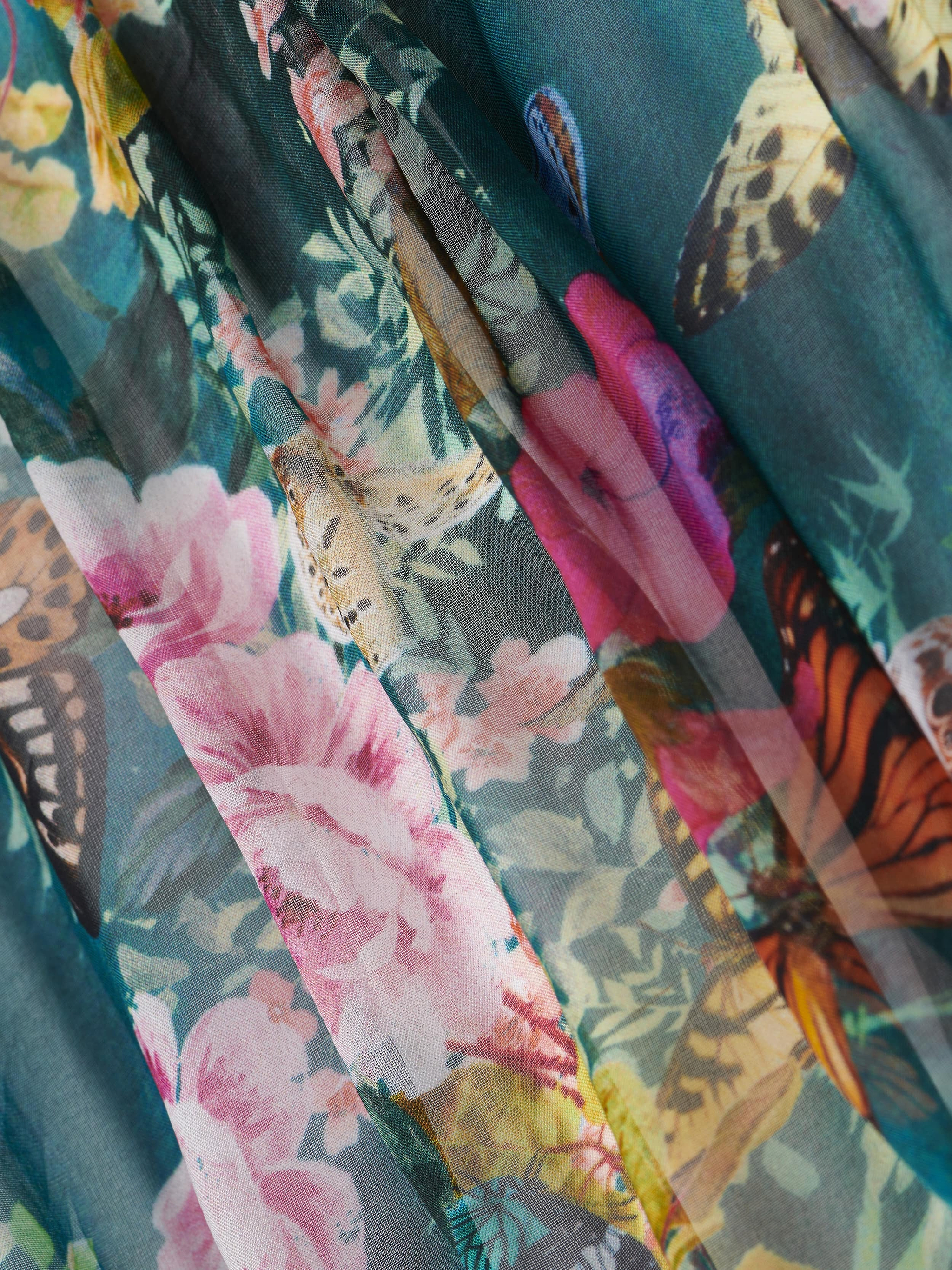 Close-up of the Sharon Cover-Up Pant, showcasing colorful floral and butterfly patterns on semi-sheer, flowing fabric beachwear with pink flowers and multicolored butterflies against a green background.