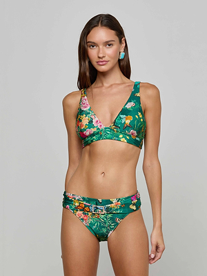 A woman with long brown hair wears the L'AGENCE Lexie Bikini Top, a green butterfly floral print V-neck bikini, with belted bottoms, standing against a plain light gray background.