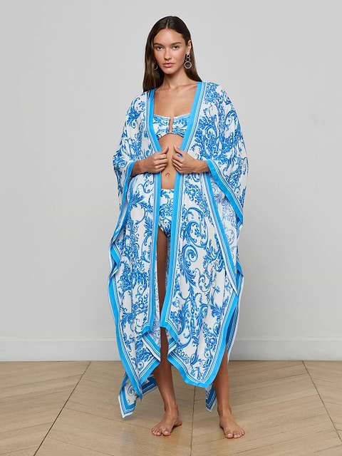 A woman stands barefoot against a plain white background, wearing the Kara Kimono Cover-Up—a long, flowing blue baroque cover-up with loose sleeves layered over a swimsuit. Crafted from water-safe viscose crepe, this elegant beachwear combines style and comfort for your next seaside getaway.