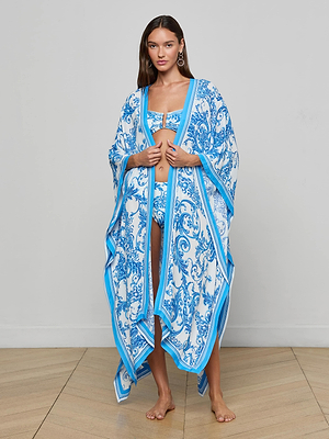 A woman stands indoors in a blue and white patterned bikini set with the Kara Kimono Cover-Up, which features an asymmetric hem. She has long brown hair, dangling earrings, and is barefoot on a light wooden floor against a plain wall.
