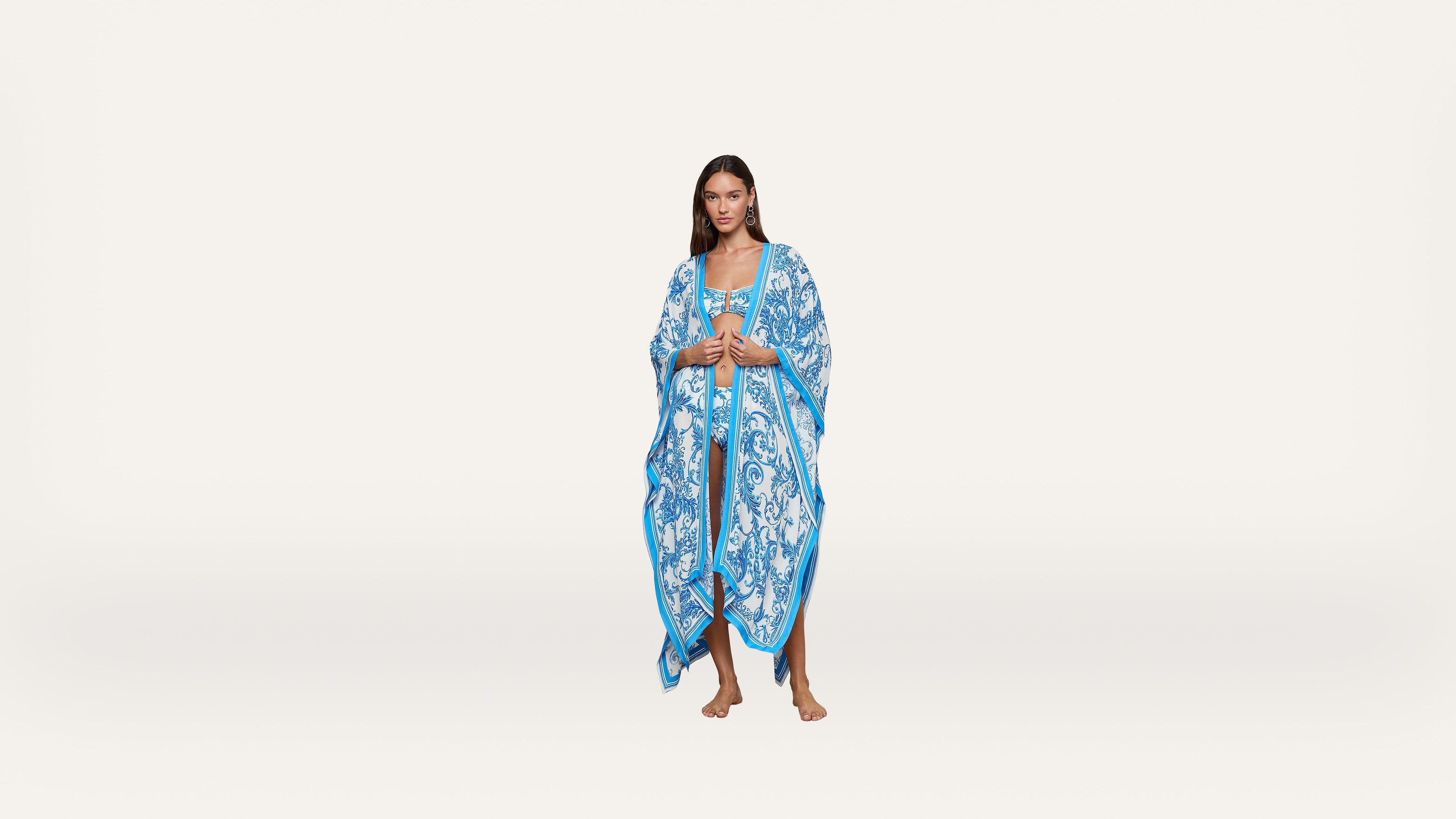 A woman stands barefoot wearing a long, flowing blue and white kaftan with intricate patterns. The background is plain white.