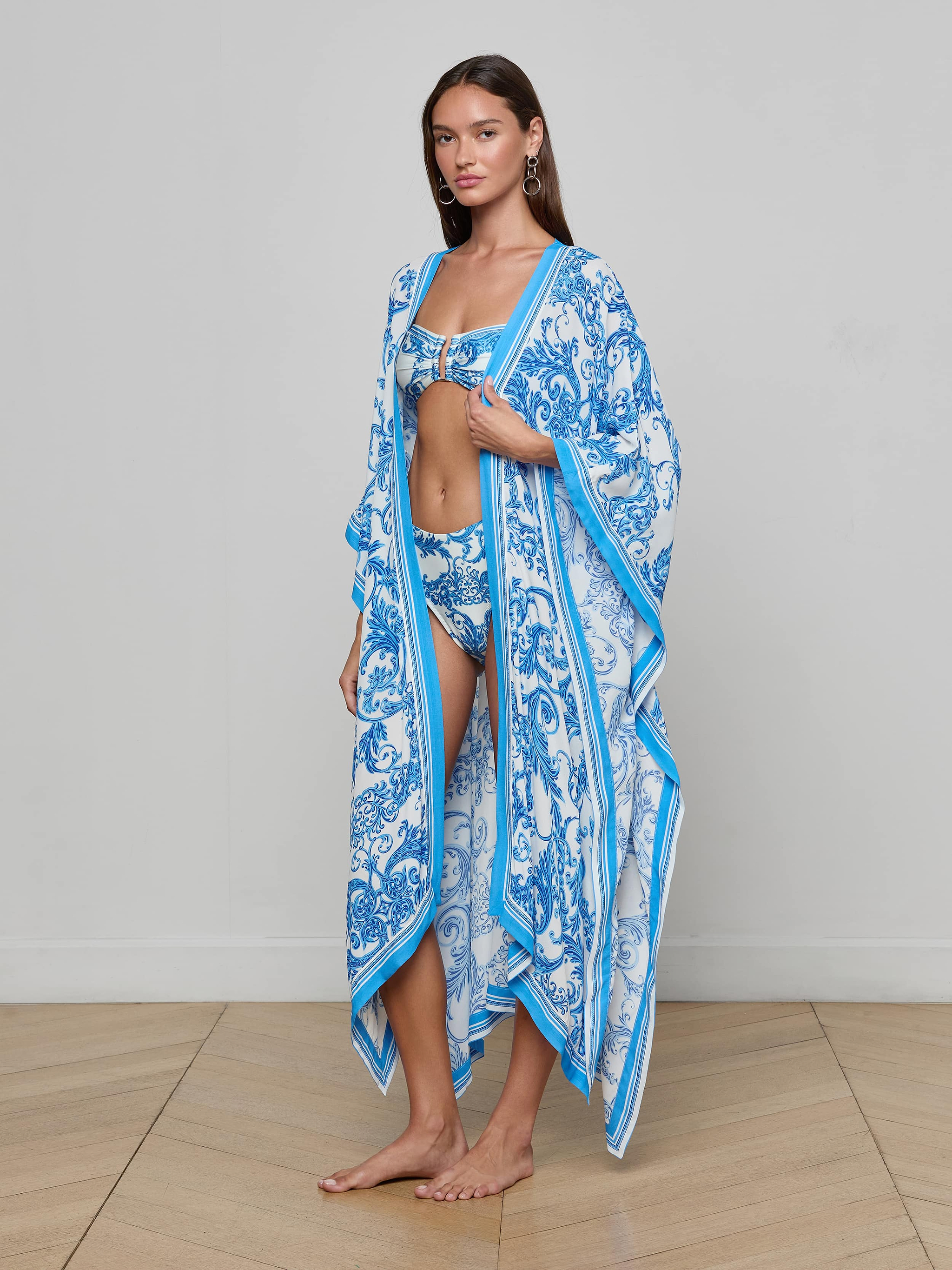 A woman stands barefoot, posing against a plain white background in a matching blue and white patterned bikini and the Kara Kimono Cover-Up—a long, flowing blue baroque piece.