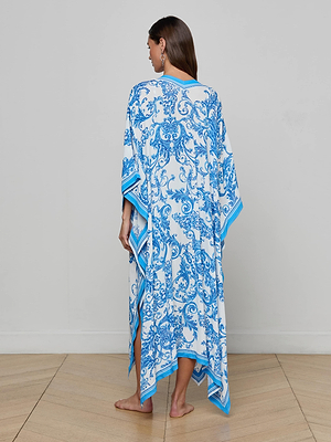 A woman with long brown hair stands barefoot on a wooden floor, facing away from the camera, wearing the Kara Kimono Cover-Up with blue floral patterns and an asymmetric hem, against a plain light-colored wall.