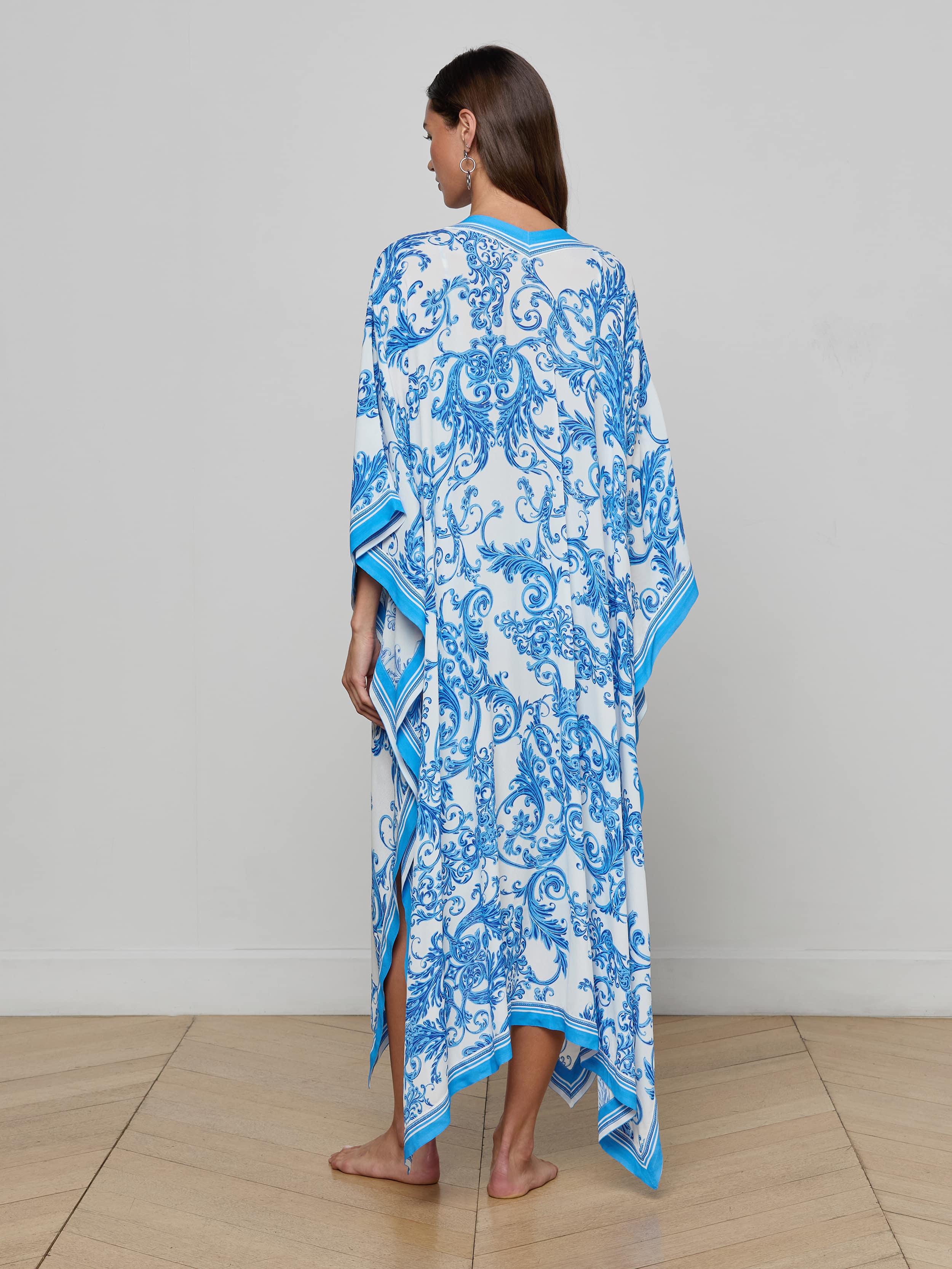 A woman stands barefoot, facing away, wearing the Kara Kimono Cover-Up—a loose-fitting, white and blue patterned beachwear kaftan with short sleeves and a flowing hem, crafted from water-safe viscose crepe—against a plain white background.