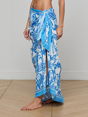 A person stands barefoot on a wooden floor, wearing the Maribel Cover-Up Pareo tied at the waist, revealing part of their leg. The plain, light-colored wall in the background adds a touch of subtle vintage glamour.