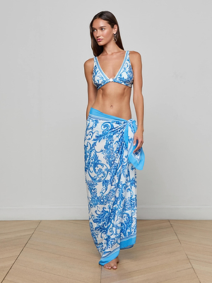 A woman poses indoors in a blue-and-white patterned bikini top and the Maribel Cover-Up Pareo styled as a skirt dress, standing barefoot on a light wood floor against a plain gray wall, radiating vintage glamour.
