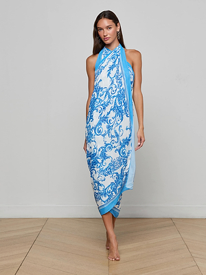 A woman stands barefoot indoors, wearing the Maribel Cover-Up Pareo, a blue halter dress with a flowing baroque design. She has long brown hair and minimal makeup, posed against a plain light gray wall and wooden floor.