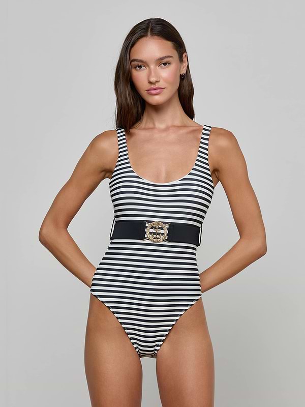 A woman stands with hands on hips against a plain background, wearing the Mila One-Piece Swimsuit—a black and white striped style featuring a wide belt with a decorative gold buckle and a scoop neckline.