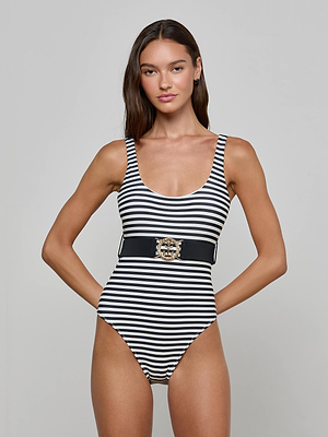 A woman with long brown hair models the Mila One-Piece Swimsuit—a black and white striped one-piece with a scoop neckline and a wide belt with gold buckle—standing against a plain background, radiating subtle French glamour.