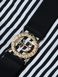 A close-up of a gold anchor belt buckle with engraved text, attached to a black strap, set against the bold black and white vertical stripes of the Mila One-Piece Swimsuit.
